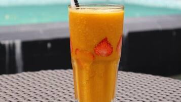 A tall glass of orange juice with a straw and a black cap, placed on a woven table with a pool and greenery in the background.