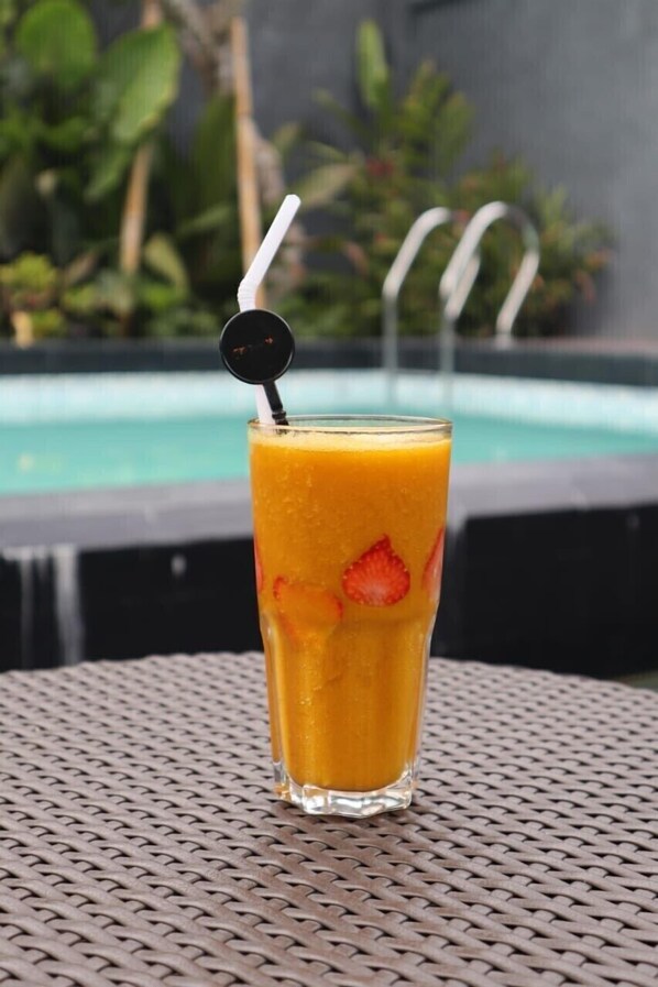 A tall glass of orange juice with a straw and a black cap, placed on a woven table with a pool and greenery in the background.