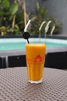 Un vaso alto de jugo de naranja con una pajita y una tapa negra, sobre una mesa de fibras naturales, con una piscina y follaje al fondo.