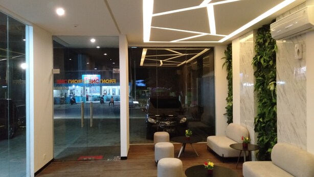 A modern lobby with a geometric ceiling design, a seating area with sofas and a coffee table, and a large window overlooking a busy street. - Front One Inn Kediri (Ngasem)