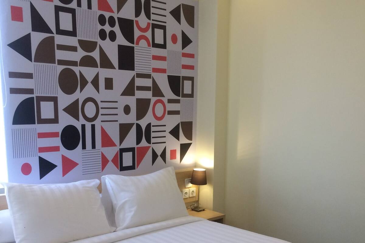 A hotel room with a bed, pillows, a nightstand with a lamp, and a geometric-patterned wall.