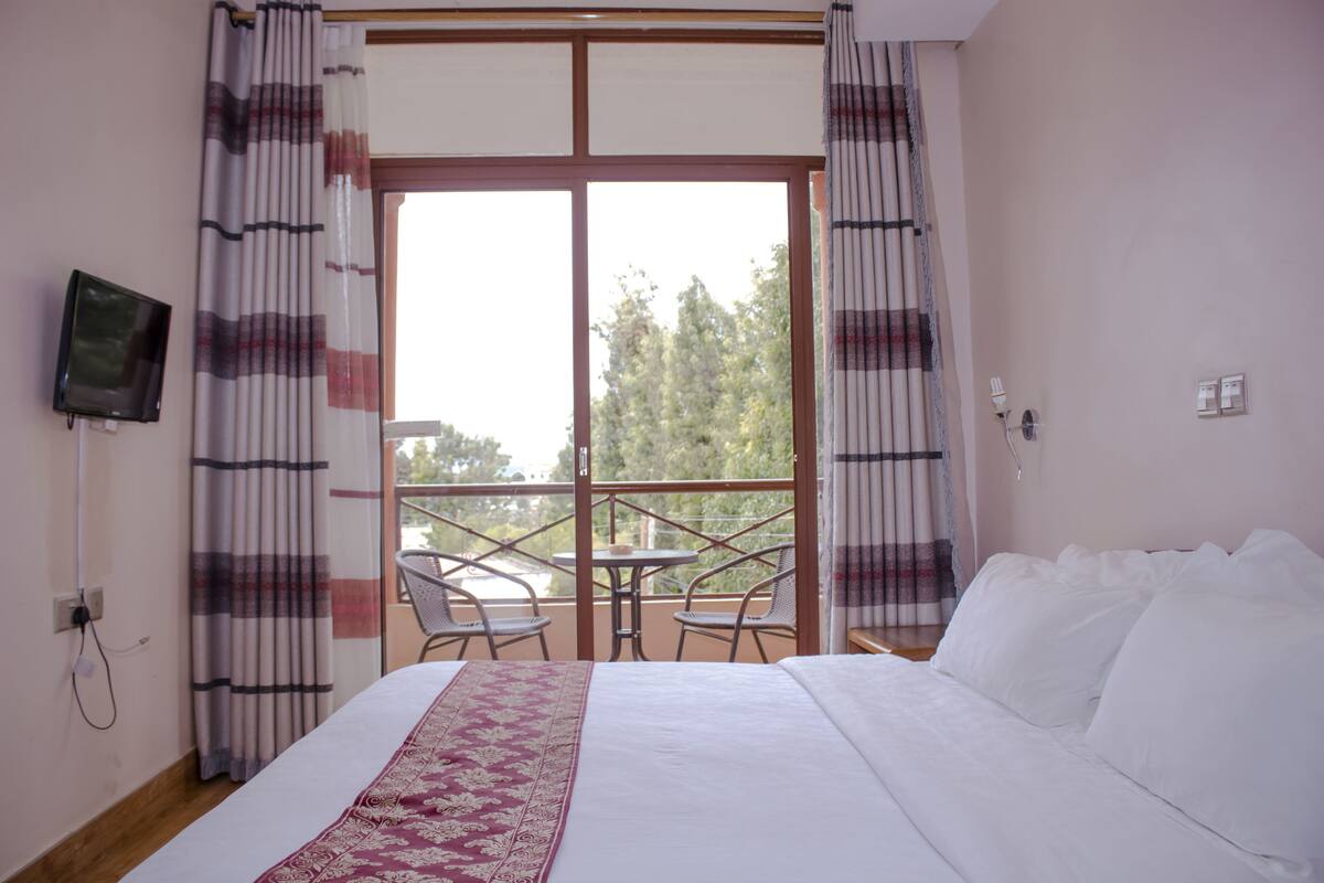 Una habitación de hotel con una cama, un televisor en la pared, un balcón con sillas y cortinas.