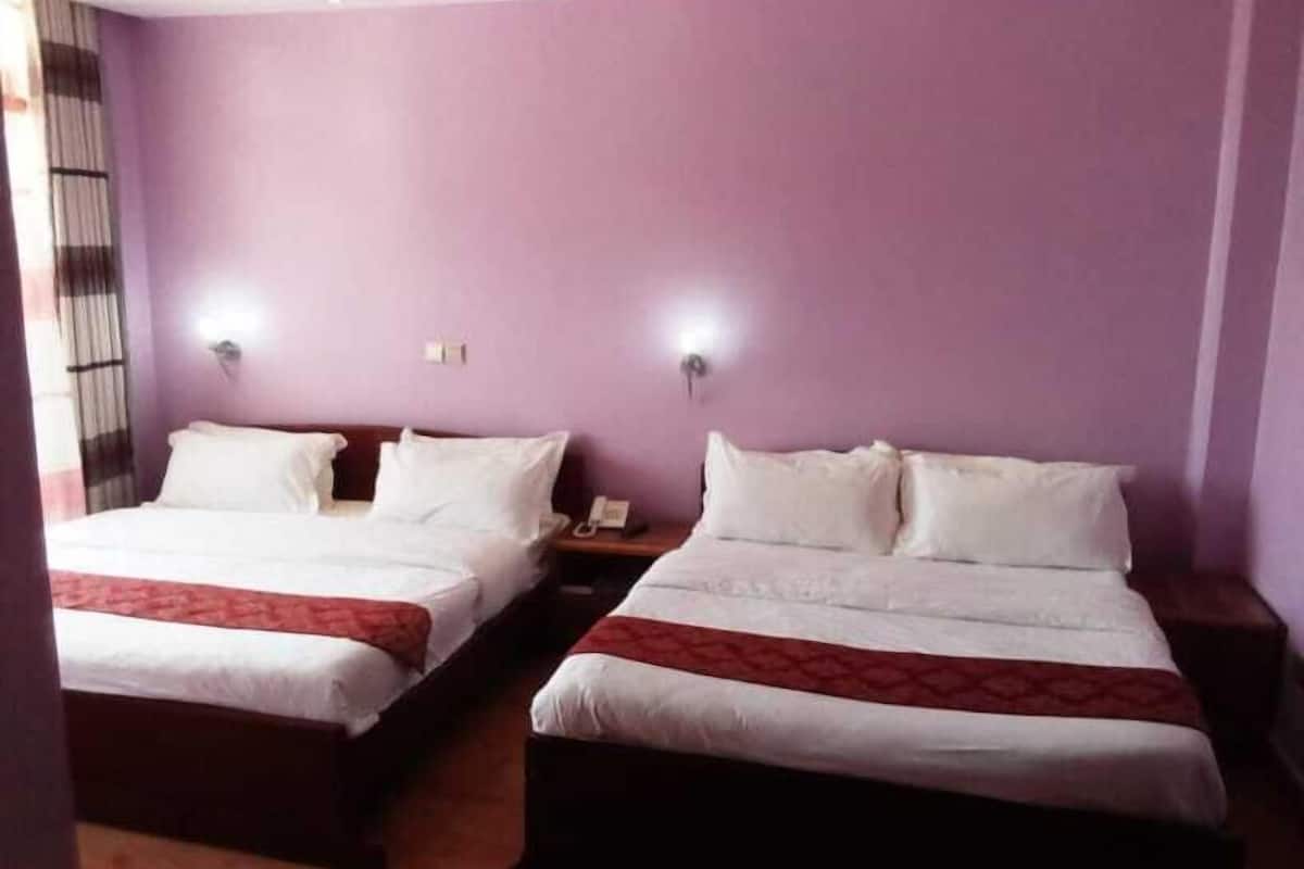 Habitación de hotel con dos camas, cada una con sábanas blancas y una colcha roja. Hay dos mesitas de noche con lámparas y un teléfono. Las paredes son moradas y hay una ventana con cortinas.