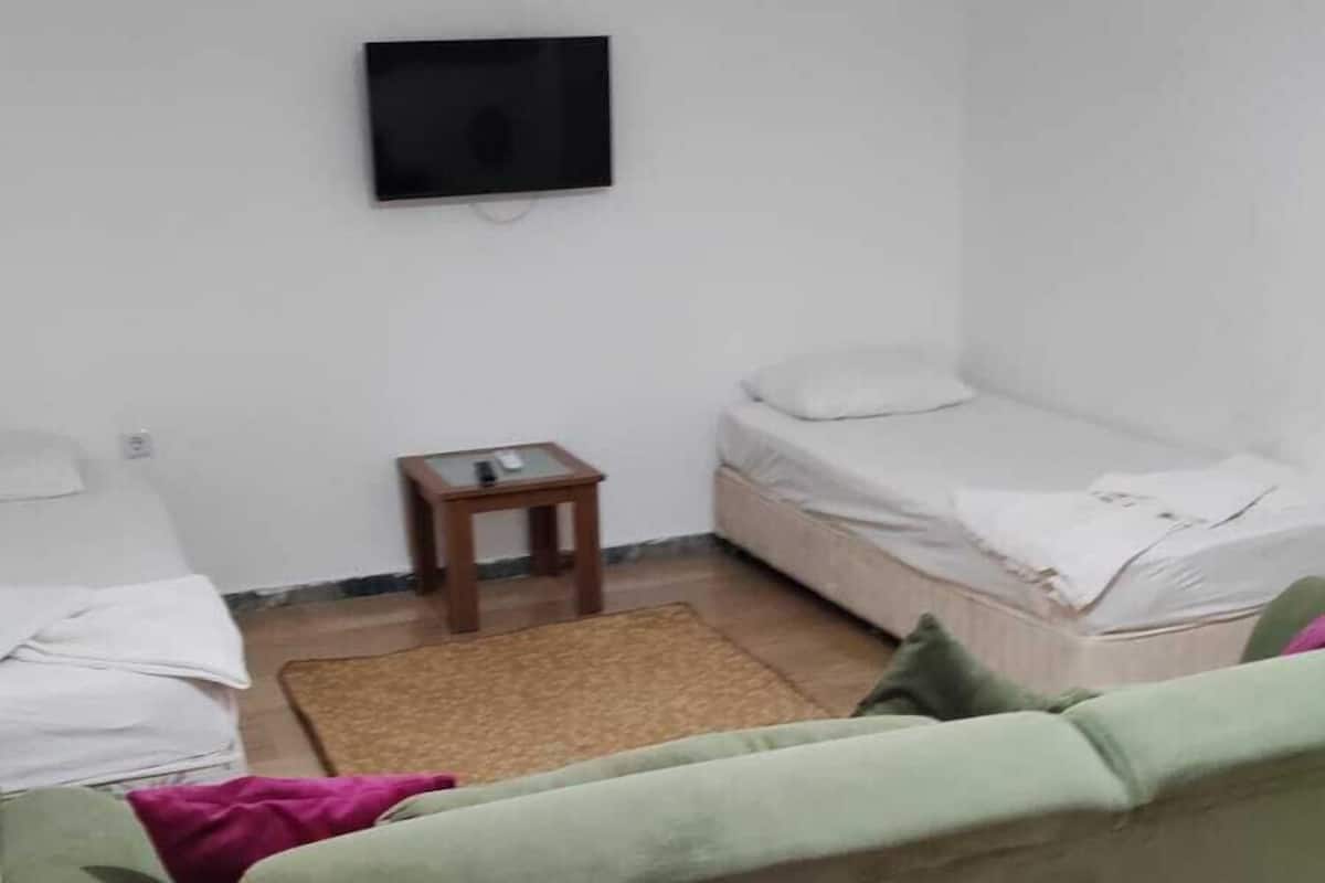 A small, sparsely furnished room with a green sofa, a small wooden table, a television mounted on the wall, and a bed with a white mattress.