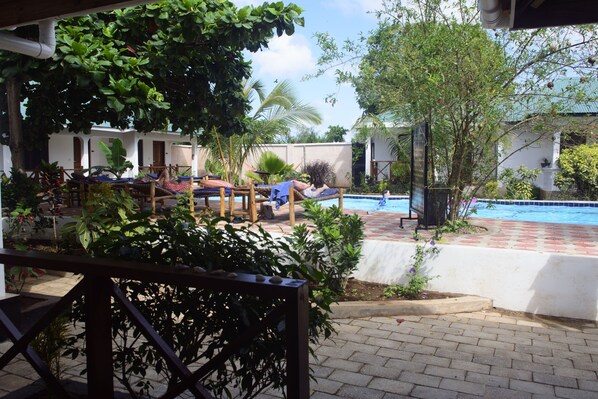 Un área de piscina con sillas de descanso y sombrillas, rodeada de follaje y un edificio blanco.