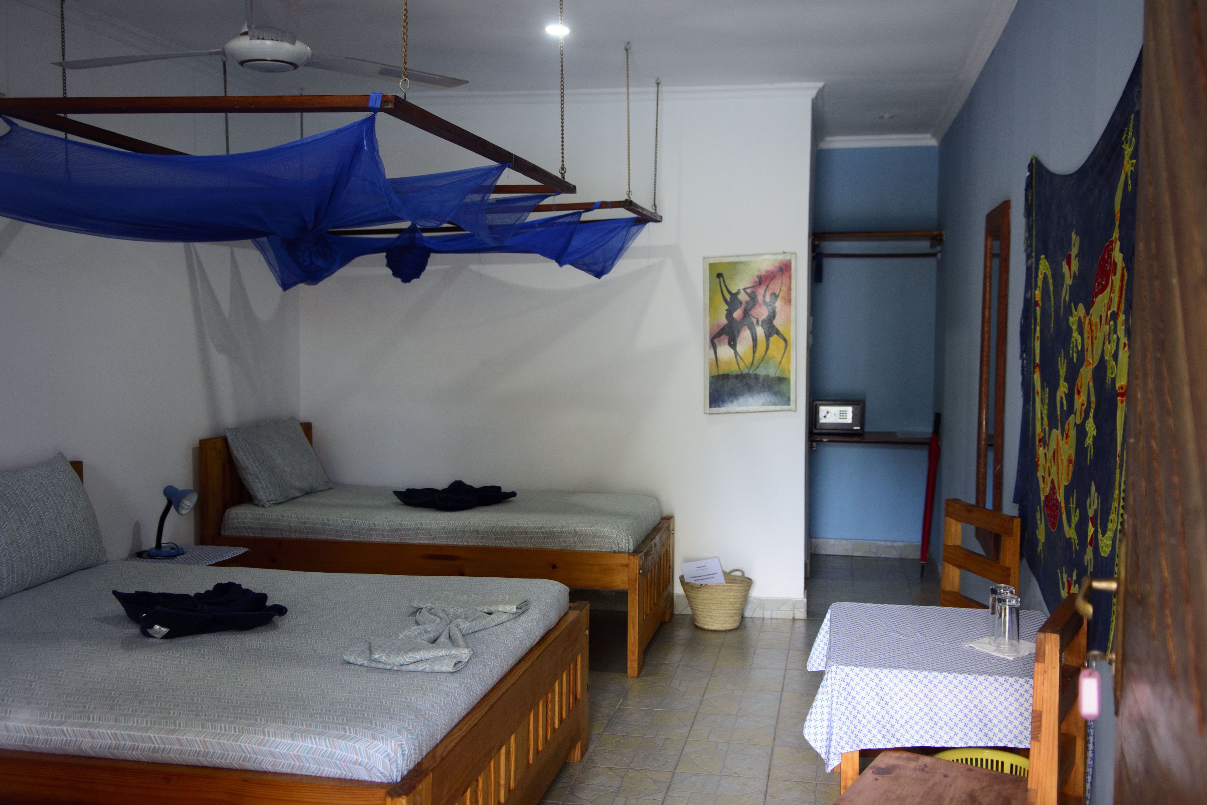 Une pièce avec deux lits en bois, un ventilateur de plafond, une table recouverte d’une nappe et un mur orné d’un tableau.
