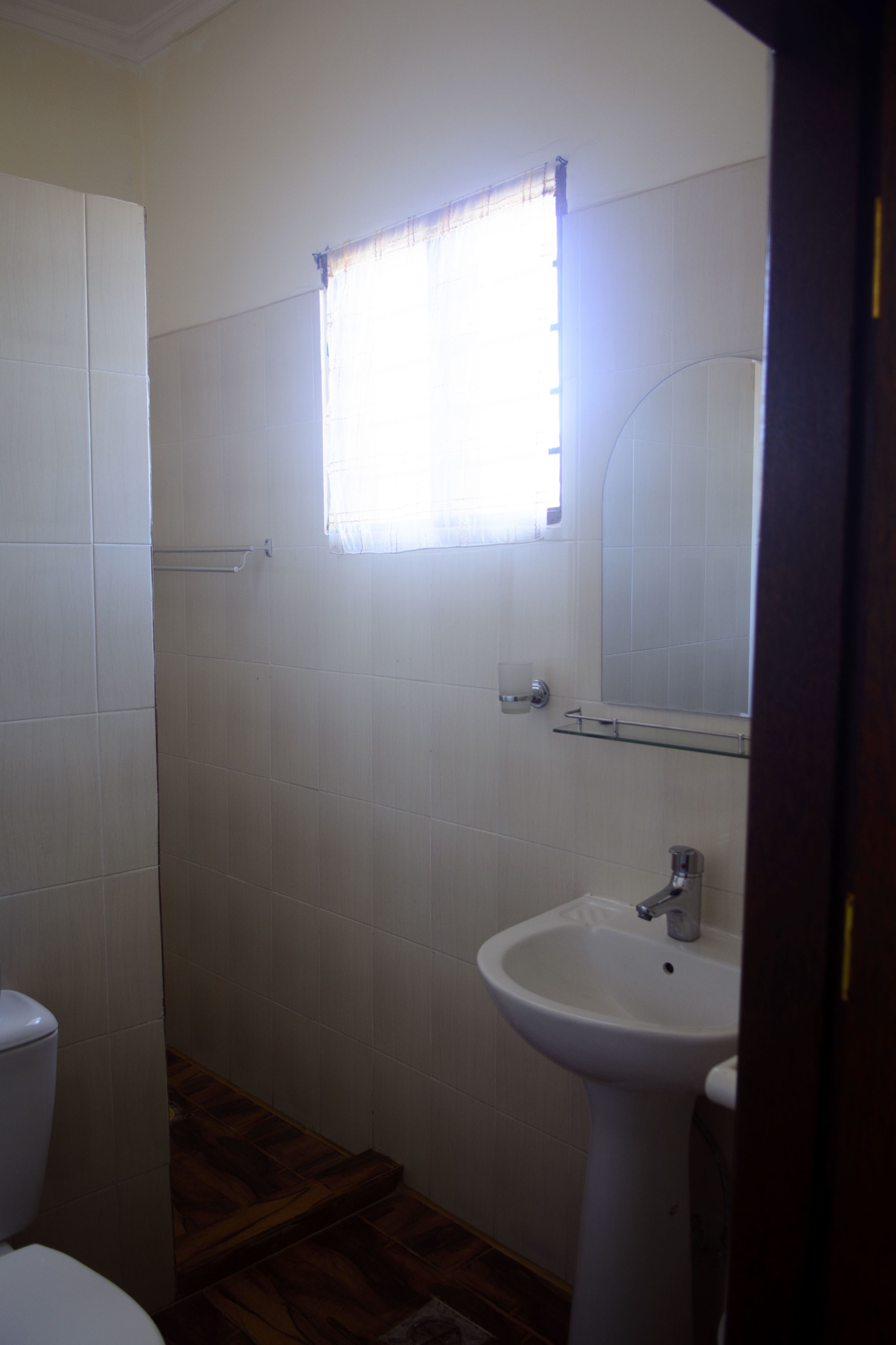 Ein Badezimmer mit weißen Fliesen, einem Fenster mit Vorhang, einem Waschbecken und einem Spiegel.