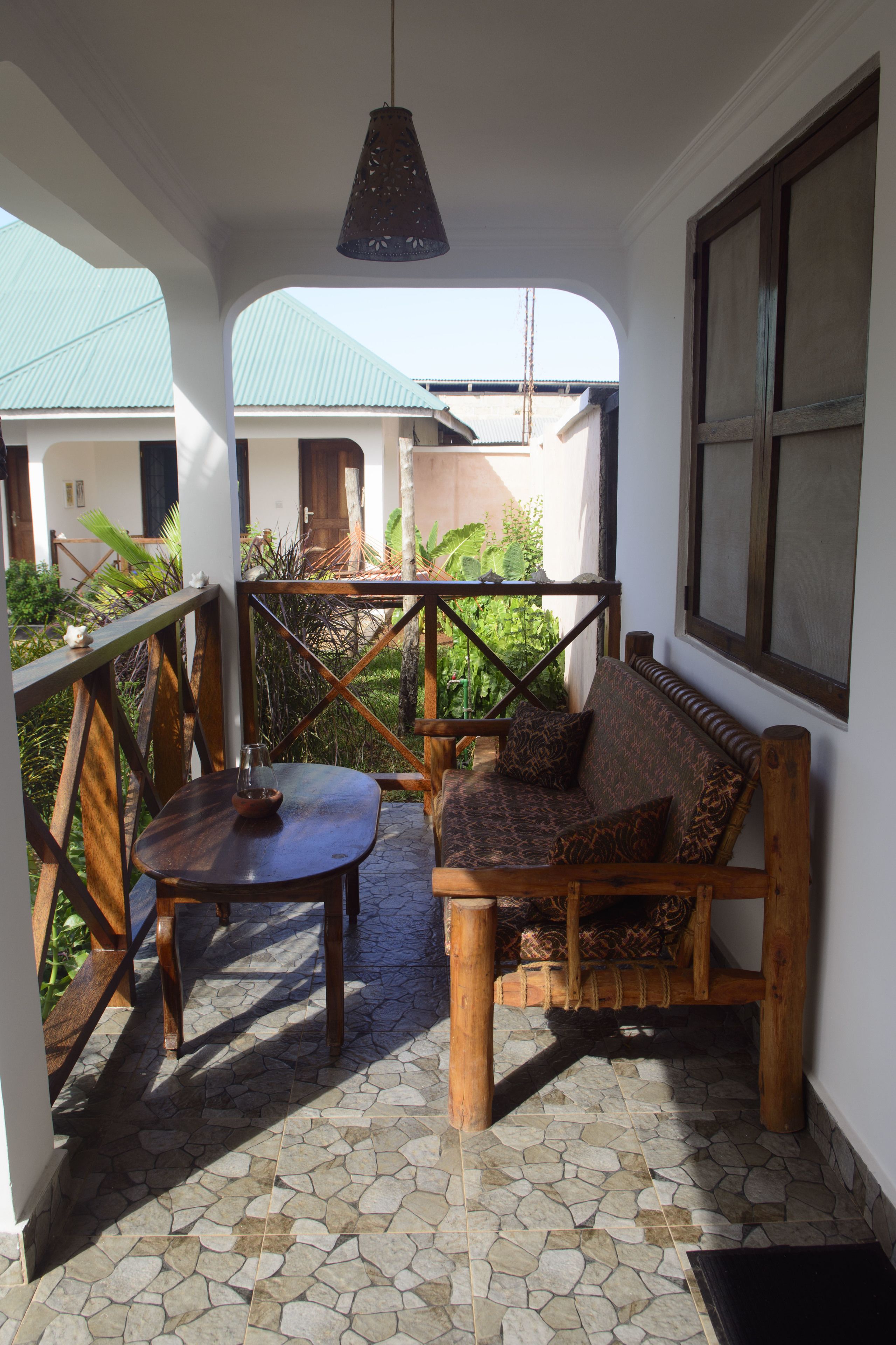 A covered outdoor seating area with a wooden bench and table, stone flooring, and a view of buildings in the background.
