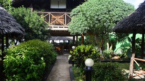 A pathway leading to a wooden deck with thatched roofs, surrounded by lush greenery and a stone floor. - Highland Bungalows (Nungwi)