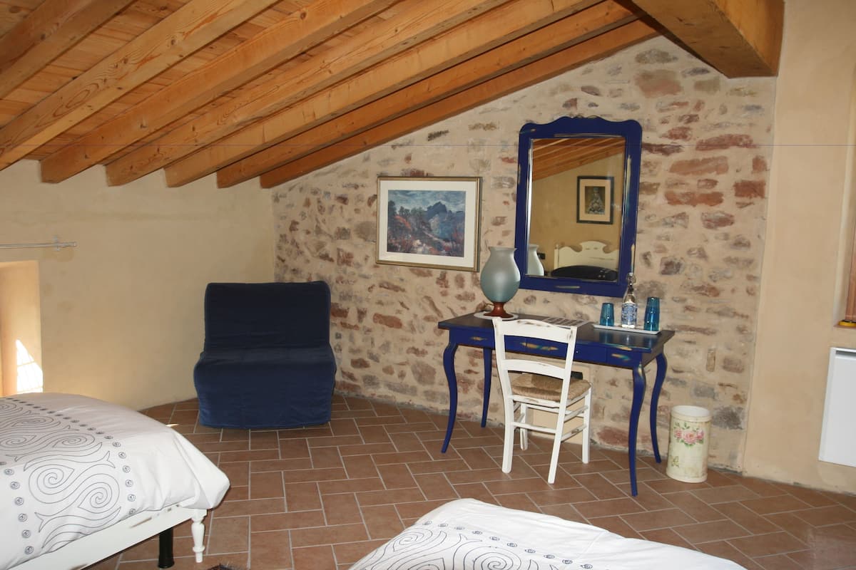 A bedroom with a wooden ceiling, a bed, a chair, a desk, a mirror, and a stone wall.
