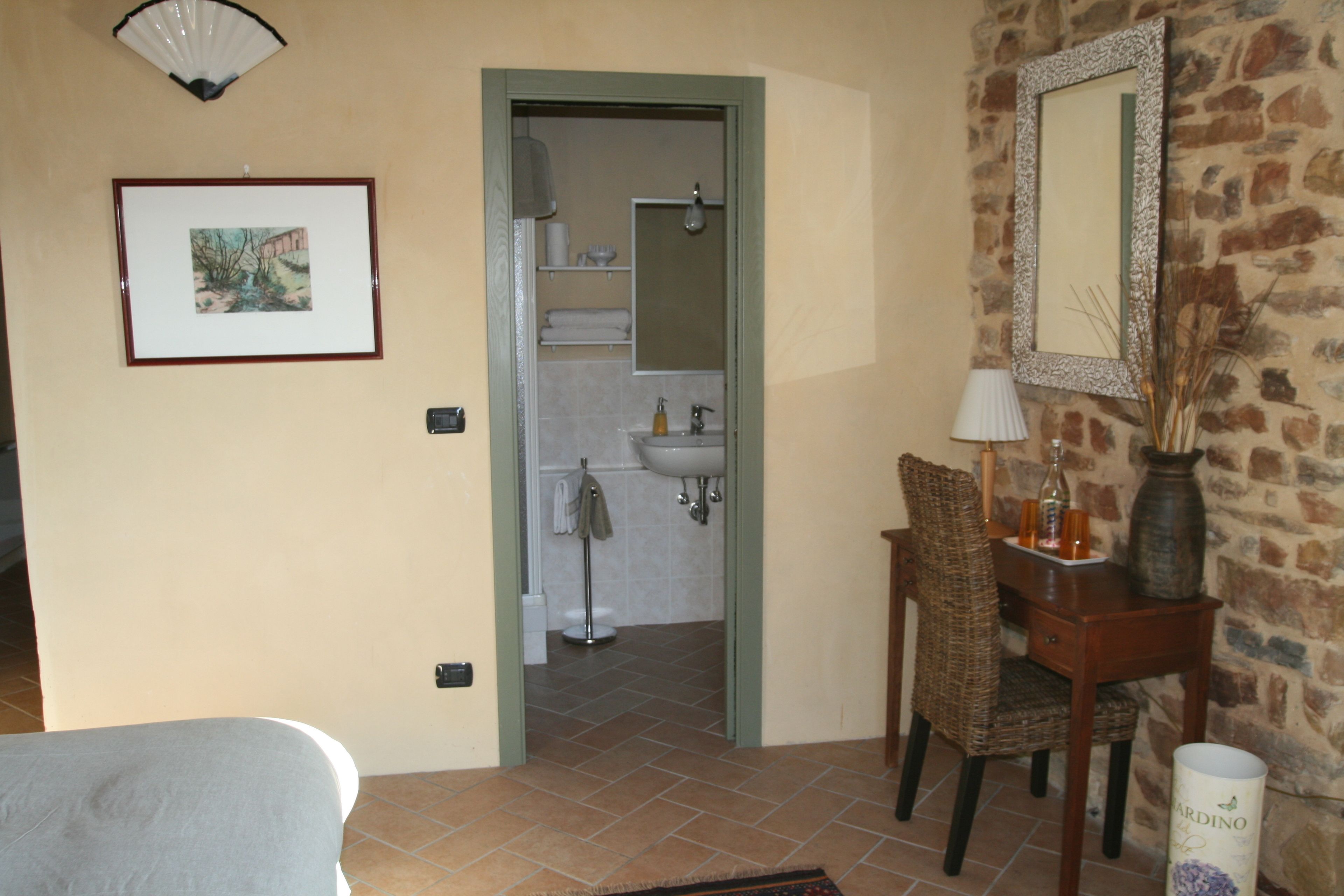Una habitació amb un llit, una cadira, una taula amb una làmpada, un vas i un quadre a la paret.
