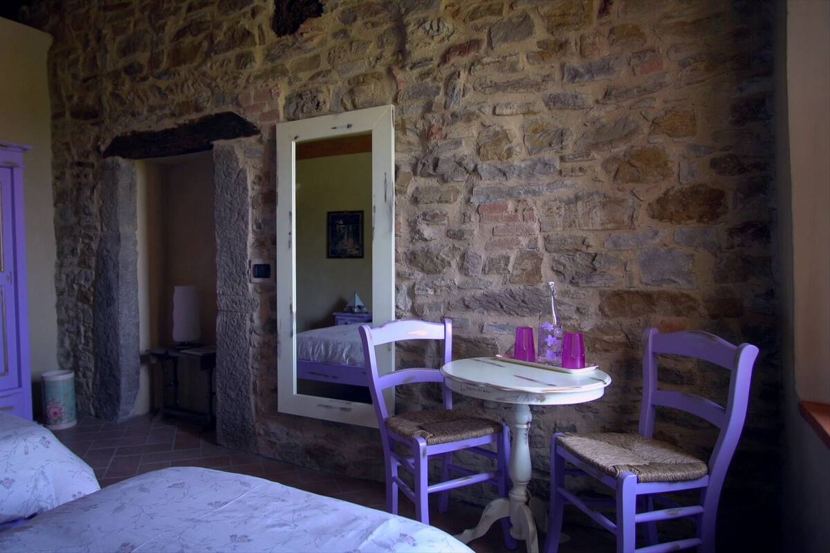 A room with a stone wall, a bed, a table with chairs, and a mirror.