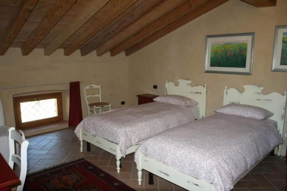 A bedroom with two beds, wooden ceiling beams, a window, and a painting on the wall.