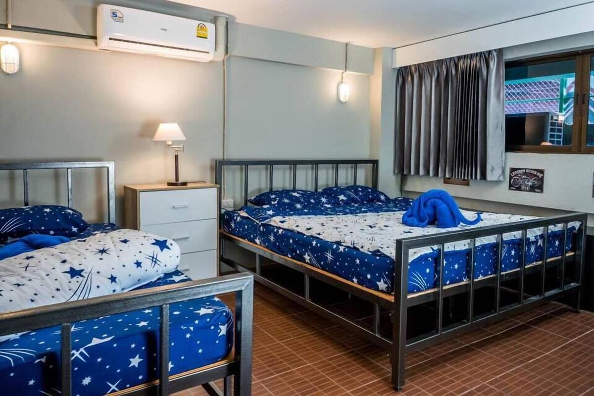 A room with two beds, each with a metal frame and star-patterned bedding. There is a small bedside table with a lamp, an air conditioning unit above the beds, and a window with curtains.