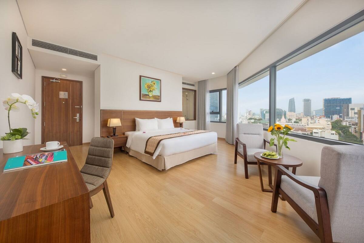A hotel room with a bed, desk, chairs, and a large window with a city view.