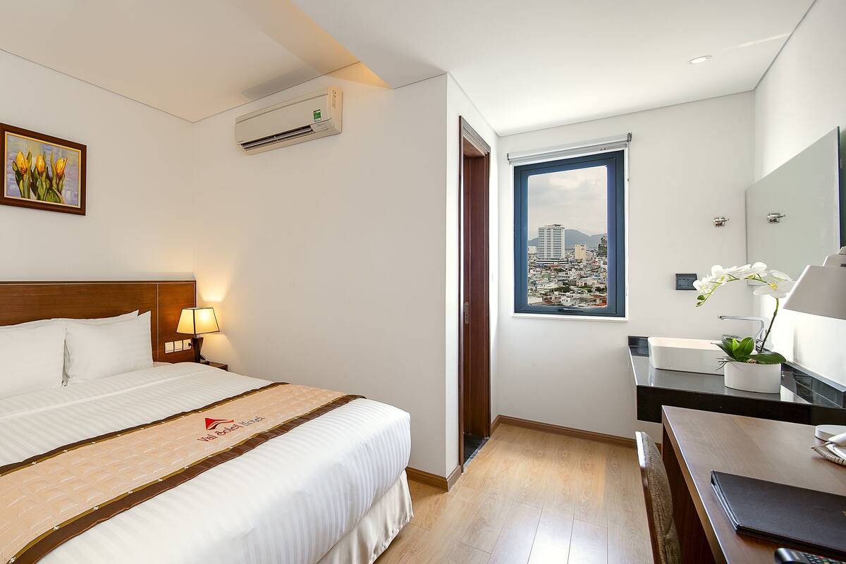 A hotel room with a bed, bedside lamps, a desk with a phone, a mirror, and a window with a city view.