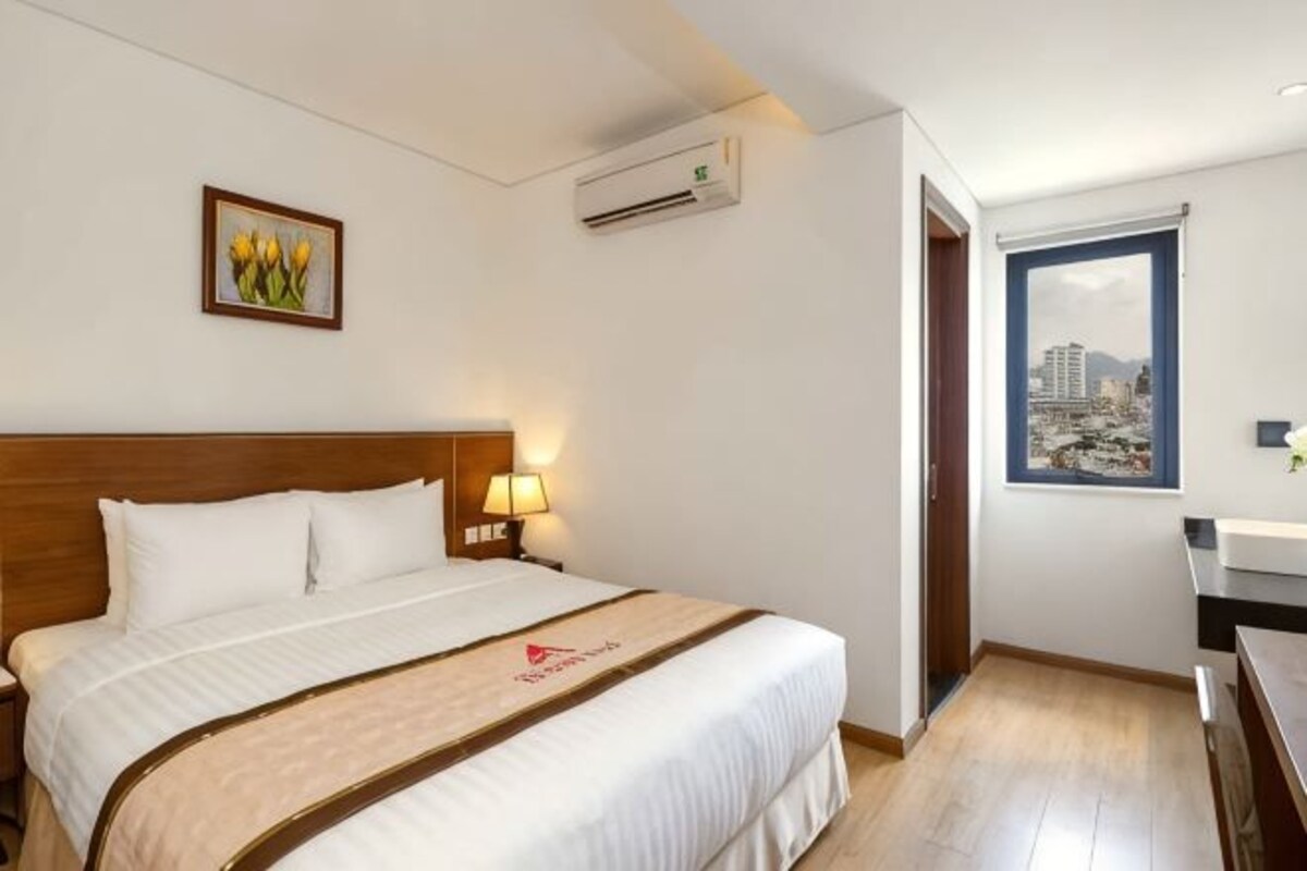 Un cuarto de hotel con una cama, una mesita de noche con una lámpara, un equipo de aire acondicionado, una ventana con vista a la ciudad y un cuadro en la pared.