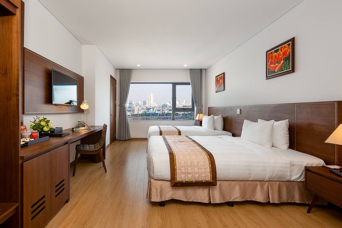 A hotel room with two beds, a desk, a chair, a TV, and a large window with a city view.