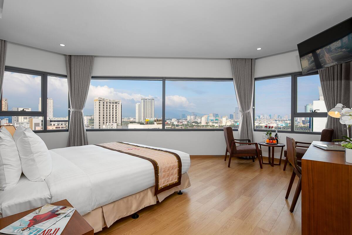 A modern hotel room with a large bed, a desk, and a view of the city skyline.