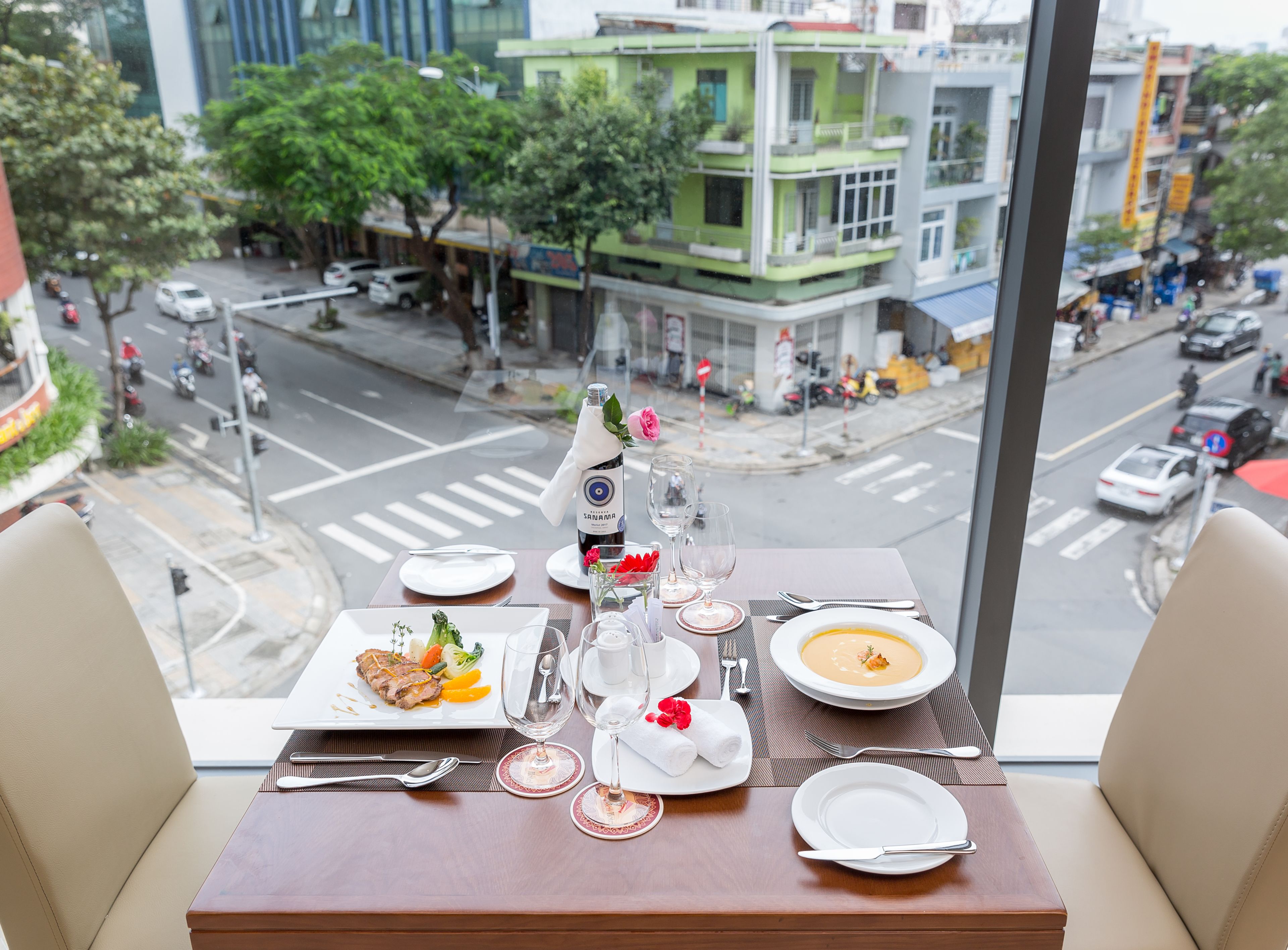 Mesa de comedor dispuesta con platos, vasos y una botella de vino Sanamaya, con vista a una calle concurrida, edificios y árboles.