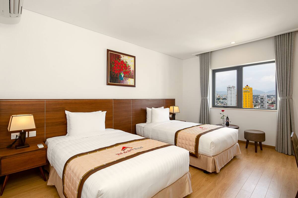 A hotel room with two beds, a desk, a chair, a window with a city view, and a painting on the wall.