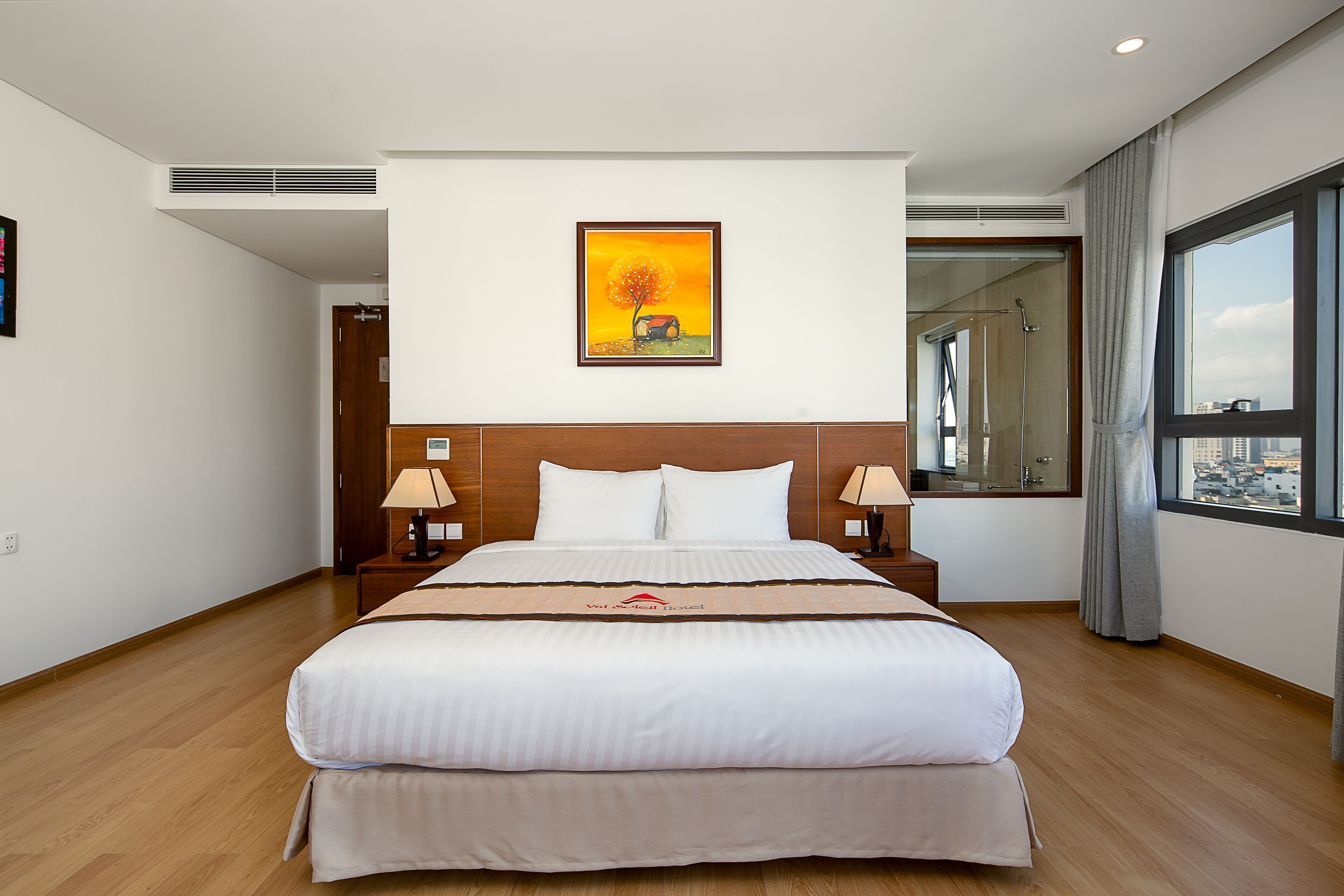 A hotel room with a large bed, a nightstand with a lamp, a painting on the wall, and a window with a view of the city.