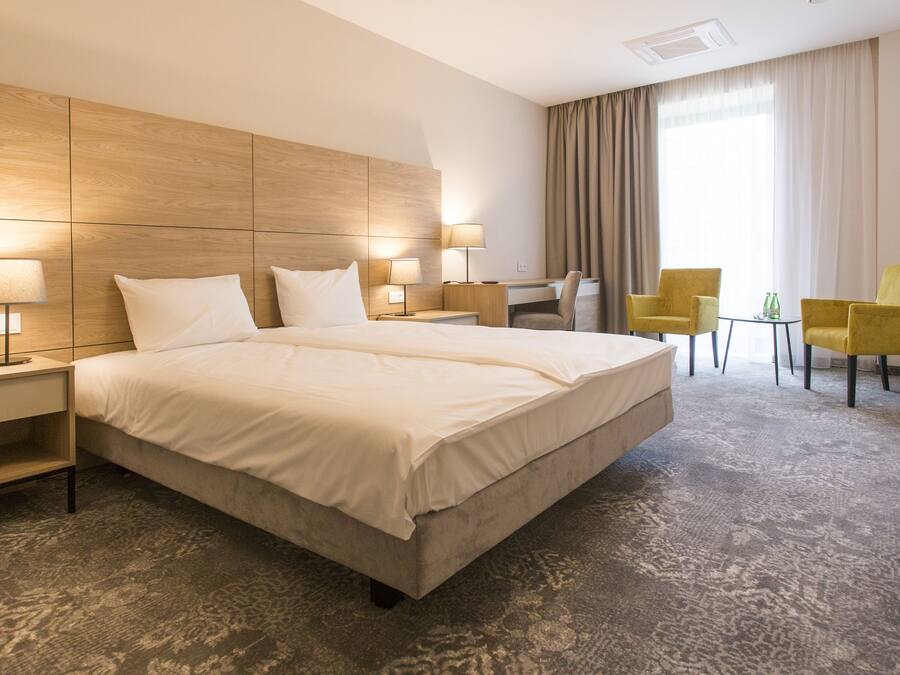 Een hotelkamer met een groot bed, twee nachtkastjes met lampen, een bureau, twee stoelen en een groot raam met gordijnen.