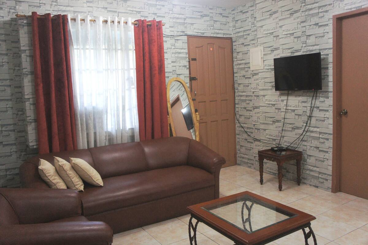 A living room with a brown leather sofa, a glass coffee table, a wooden side table, a flat-screen TV mounted on the wall, and a wooden door.