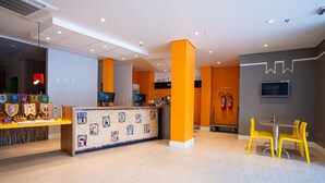 A hotel lobby with a reception desk, a children's play area, and a dining area with yellow chairs.