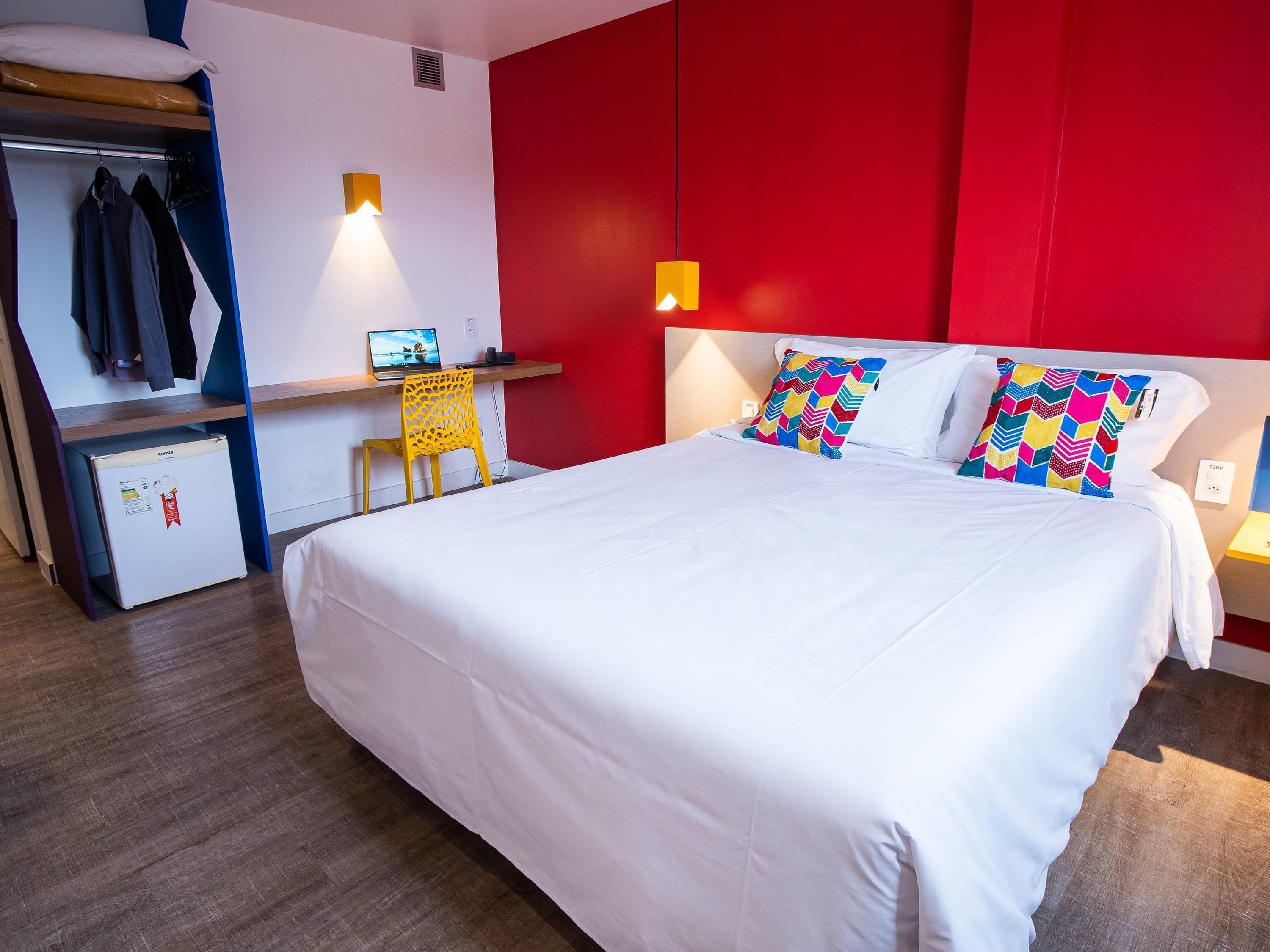 A hotel room with a bed, a desk with a computer, a small fridge, and a chair.