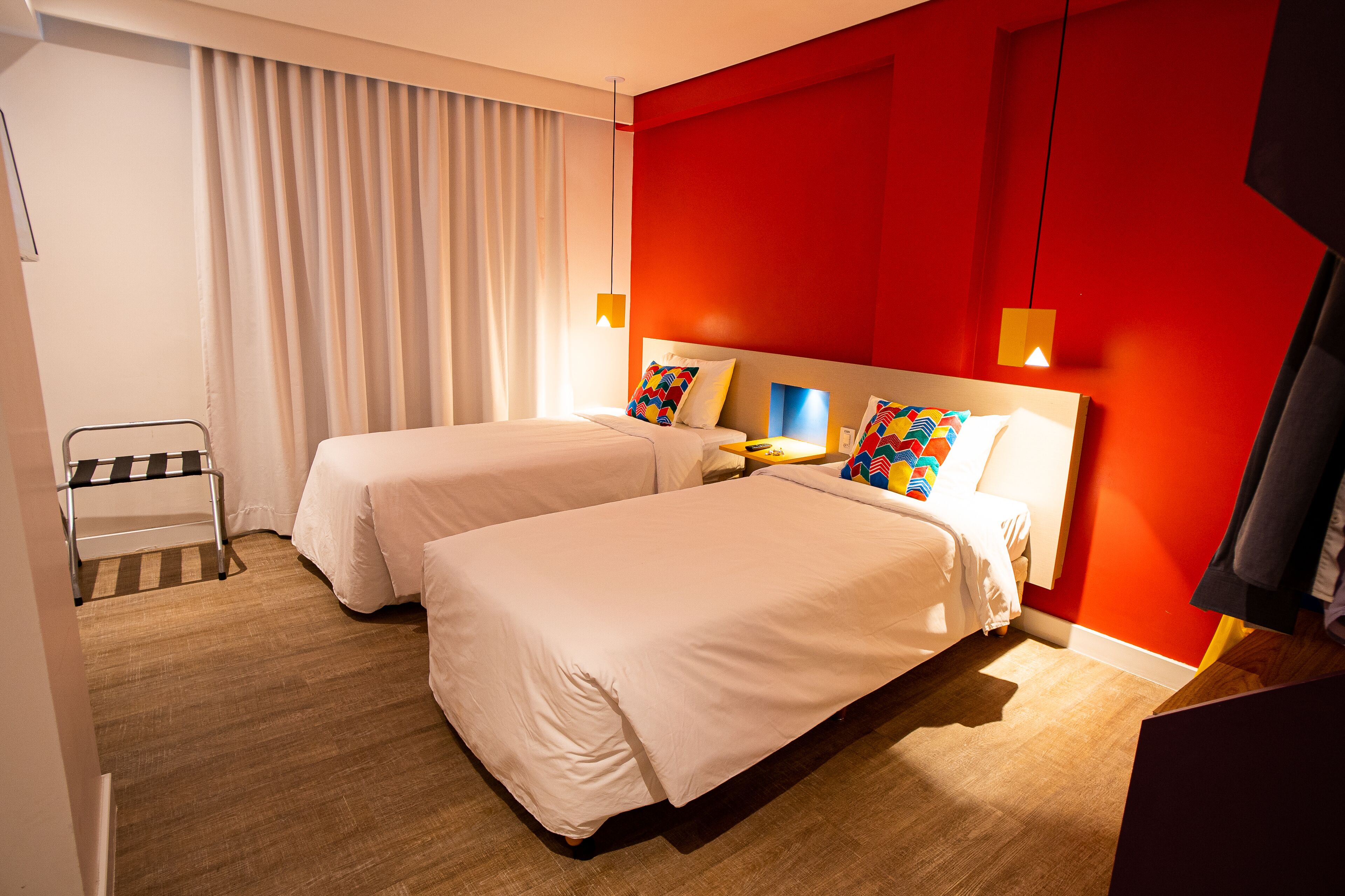A hotel room with two beds, a wooden floor, a red accent wall, and a window with curtains.
