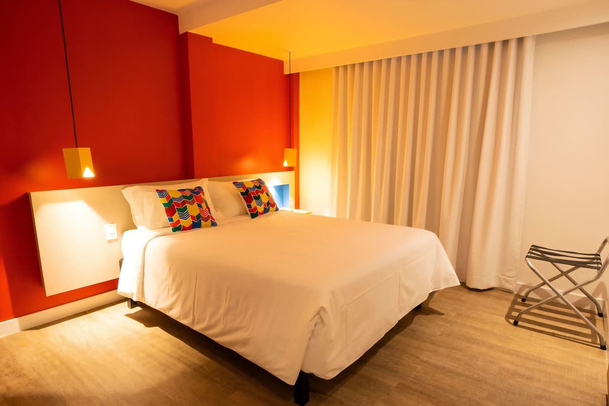 Une chambre d’hôtel avec un lit, une chaise, un rideau et un mur muni d’un luminaire.