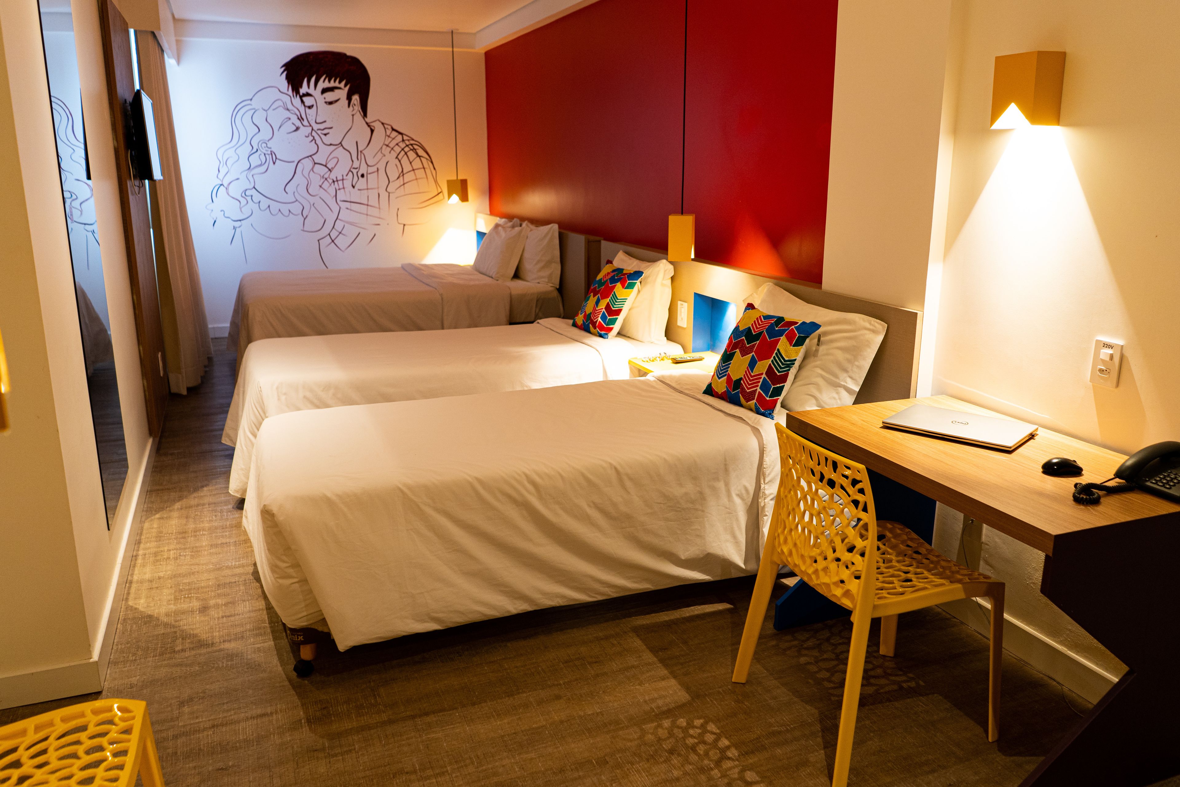 A hotel room with two beds, a desk with a chair, a wall-mounted TV, and a wall art depicting a couple.