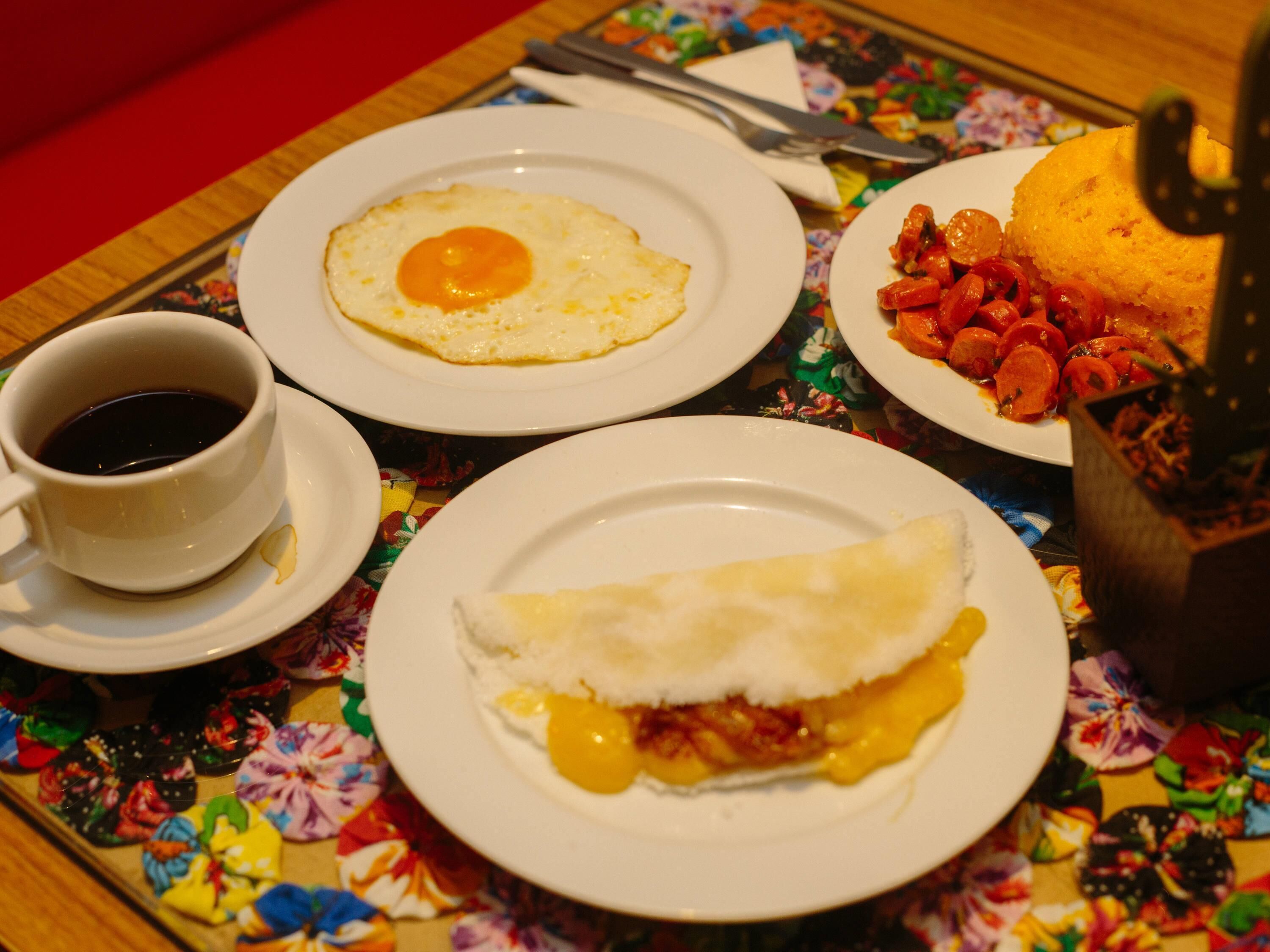 Una mesa con mantel floreado, platos de comida que incluyen una tortilla, una taza de café y un recipiente que parece contener salsa o condimento.