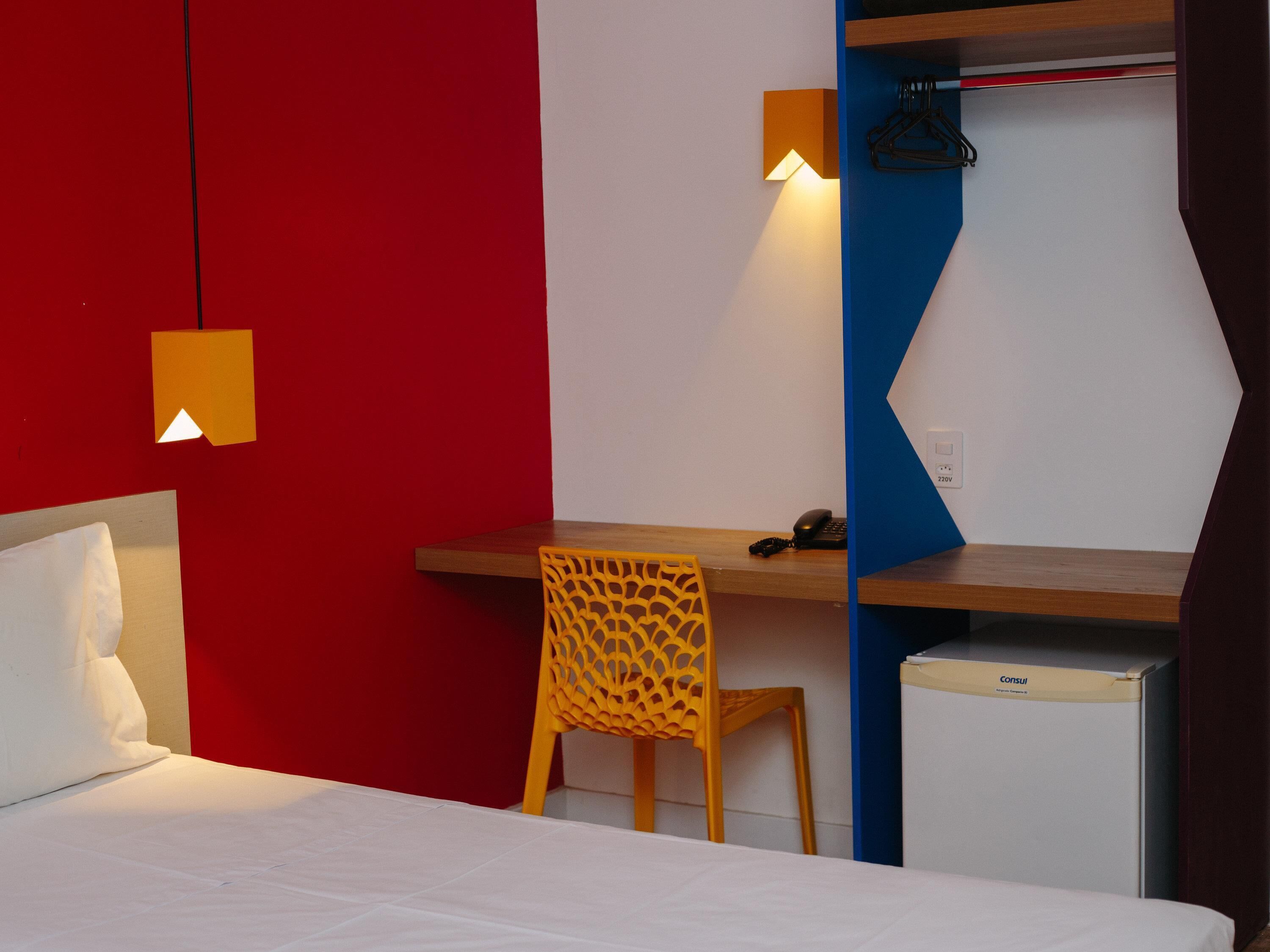 A hotel room with a bed, a desk with a chair, a small refrigerator, and a wardrobe.