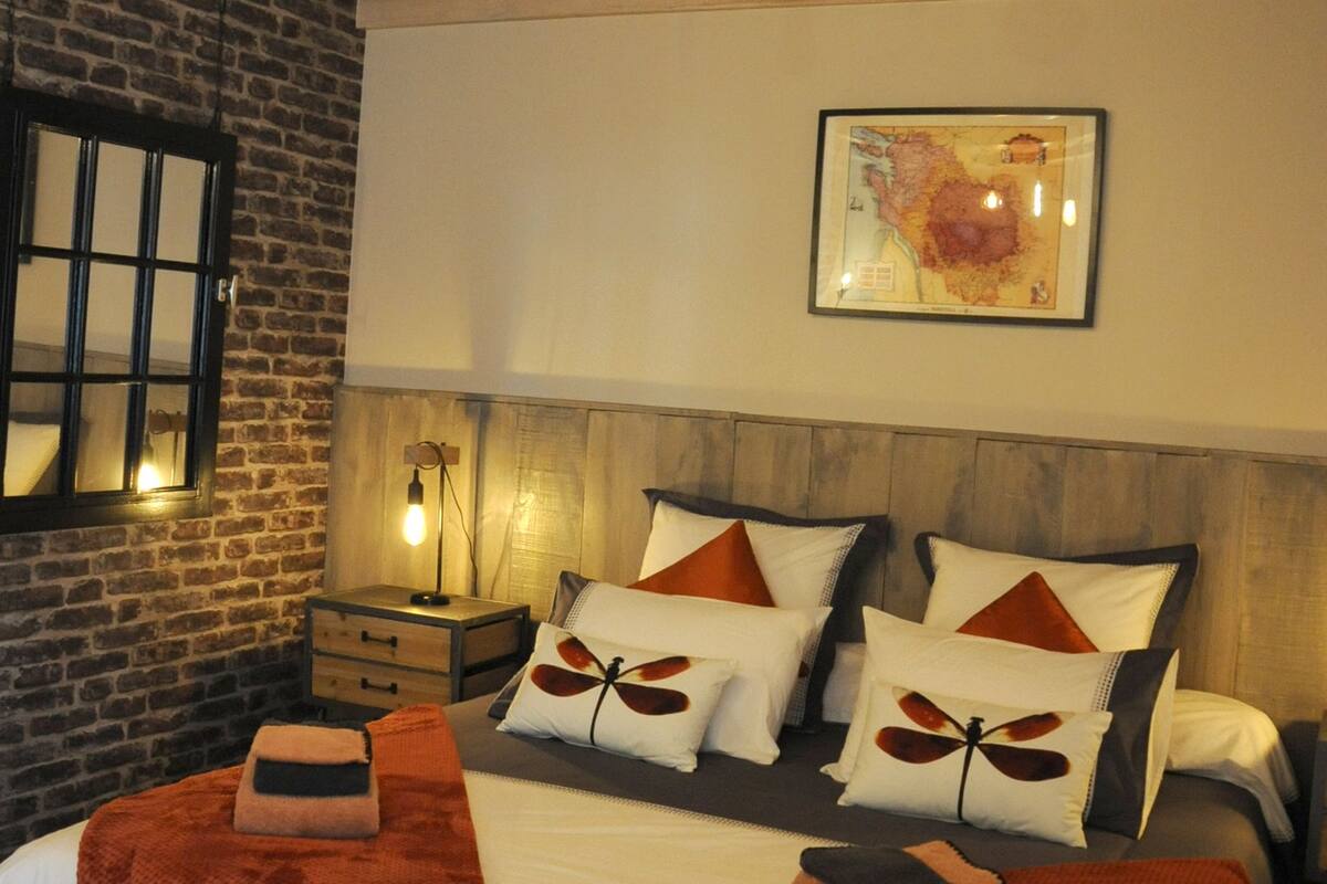Un dormitorio moderno con pared de ladrillo, techo de madera y cama con muchos almohadones y una colcha.