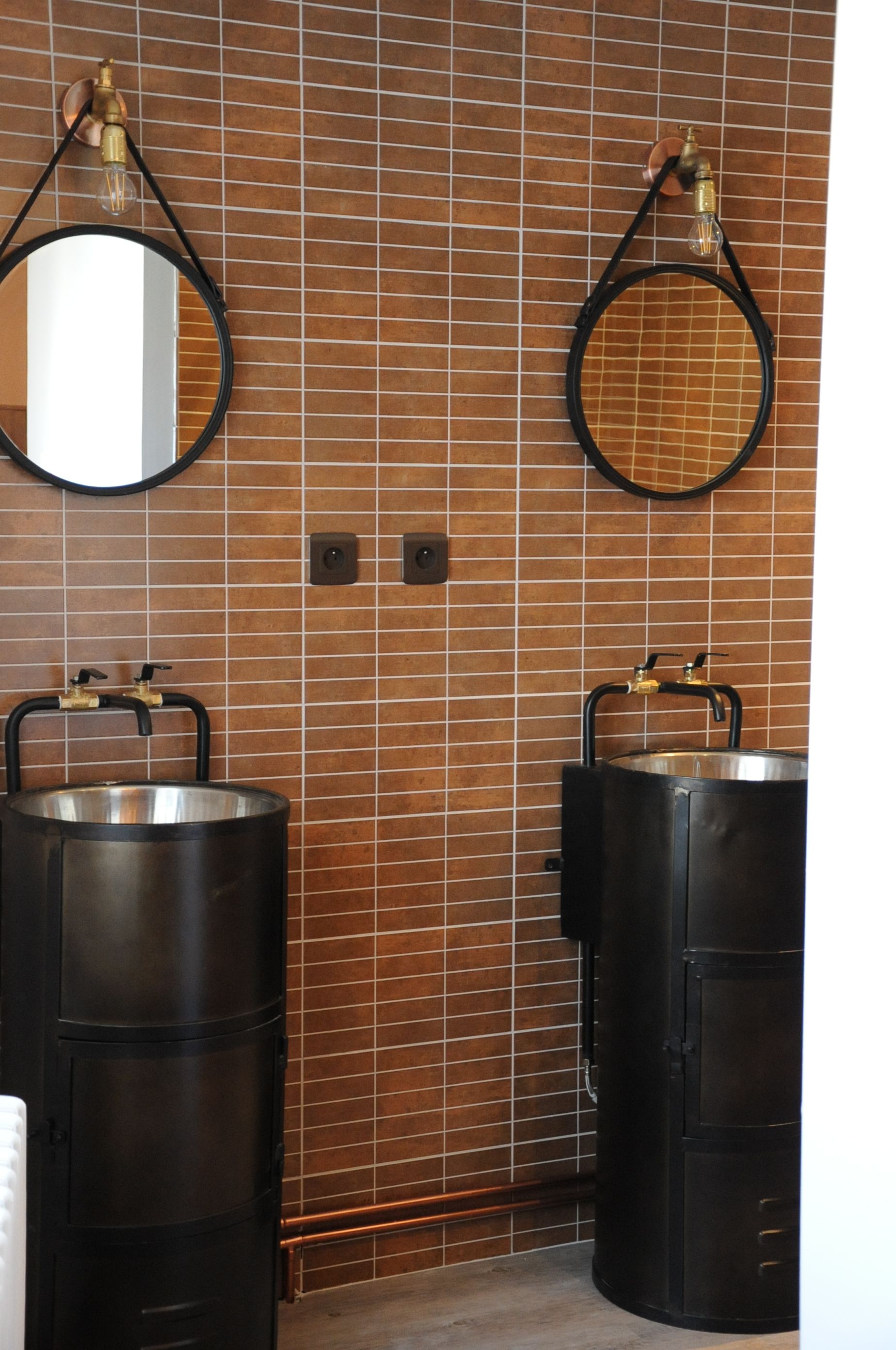 A bathroom with two sinks, each with a large metal container, a mirror, and a wall-mounted light fixture.
