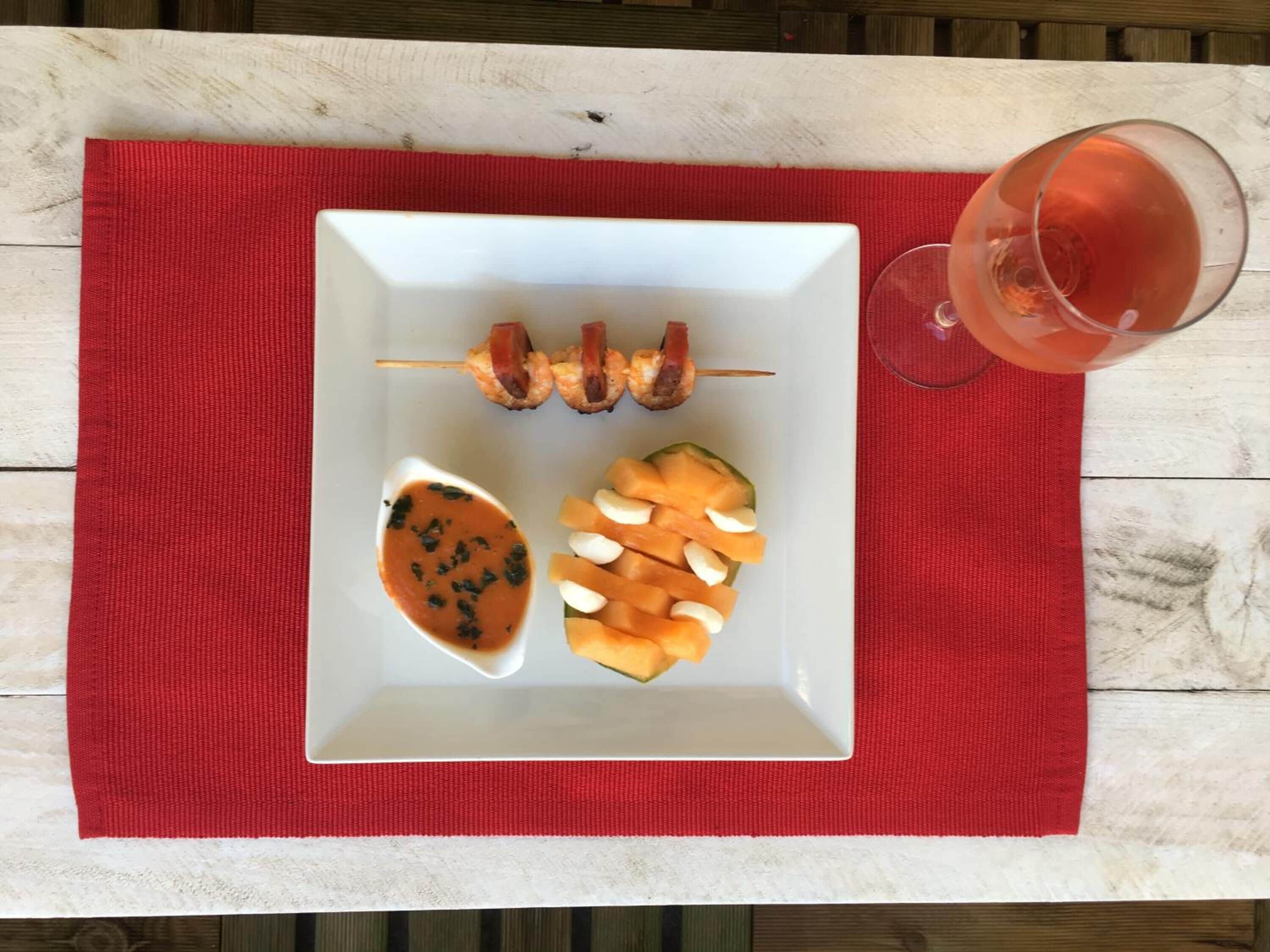 Un plato blanco con brochetas, melón y salsa sobre un tapete rojo.