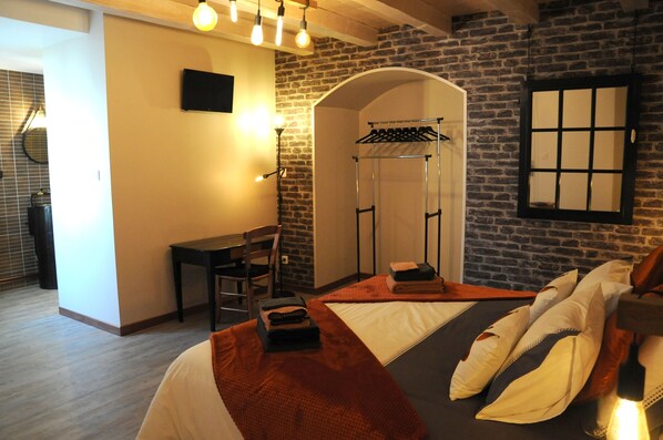 A hotel room with a bed, a desk, a chair, a hanging clothes rack, and a brick wall. - Le Nid de Sigogne (Sigogne)