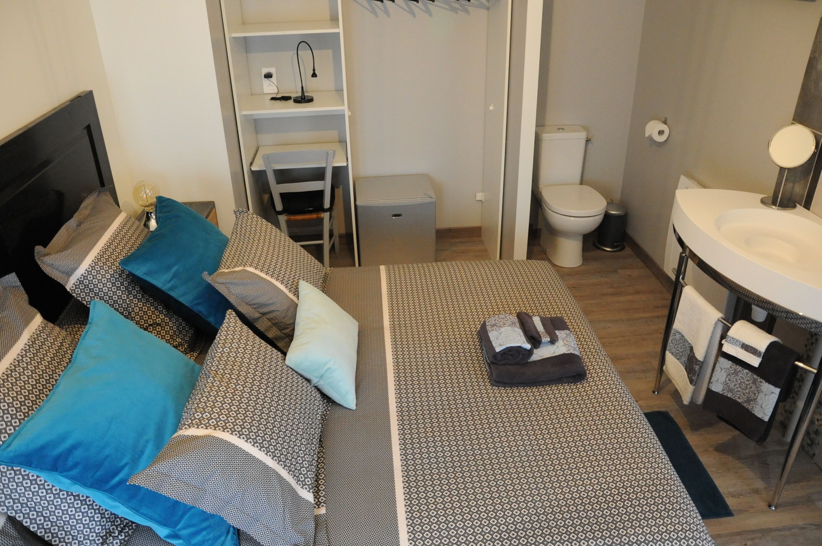Une chambre compacte comprenant un lit, des tables de chevet, un bureau, une armoire, des toilettes et une salle de bain avec un lavabo et un miroir.