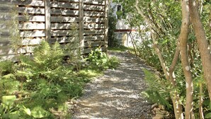 A gravel path leading to a wooden structure with a slatted roof, surrounded by lush greenery and trees. - Lille Carlsson Studio (Broby)