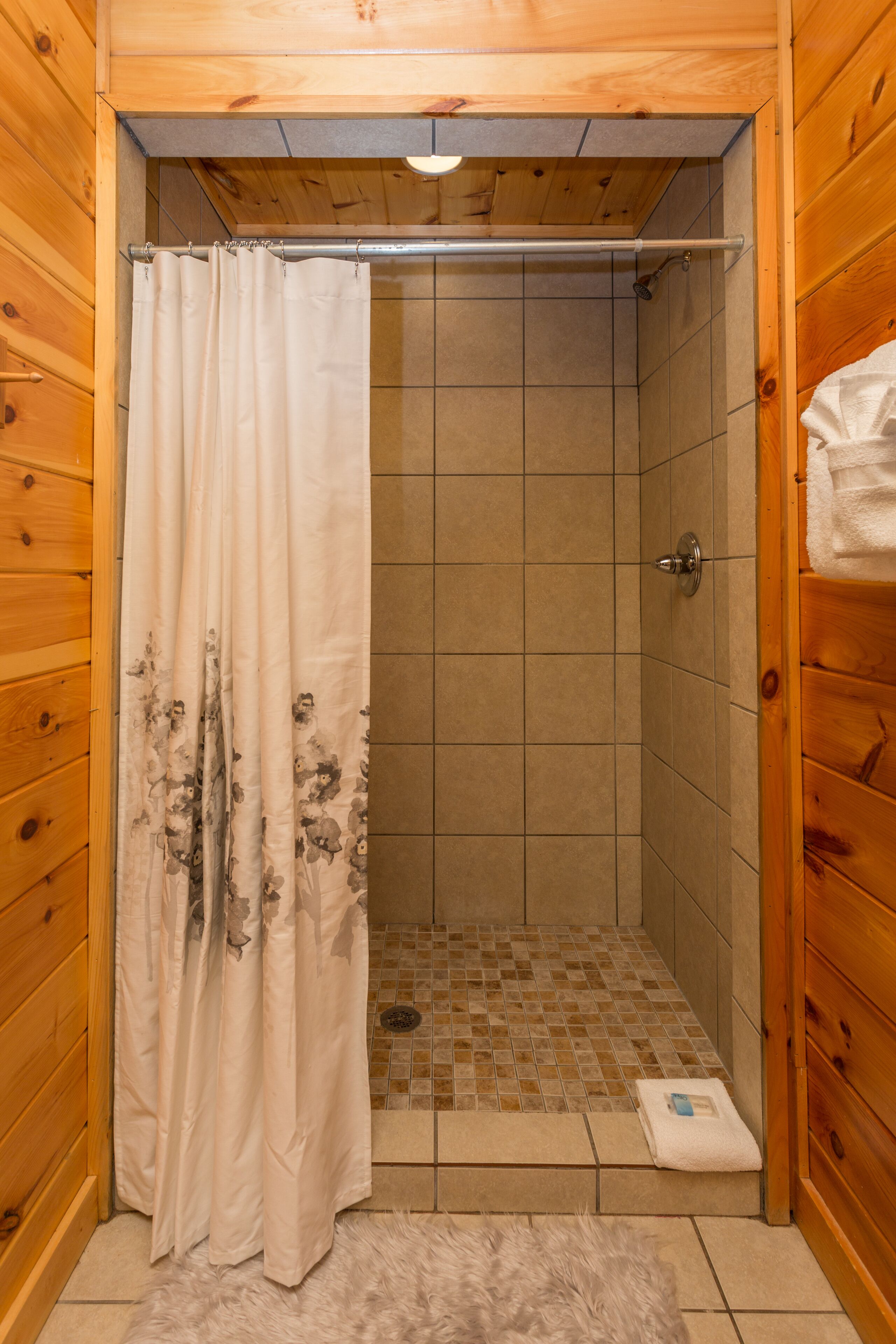 A wooden-paneled bathroom with a shower area, tiled floor, and a fluffy rug.