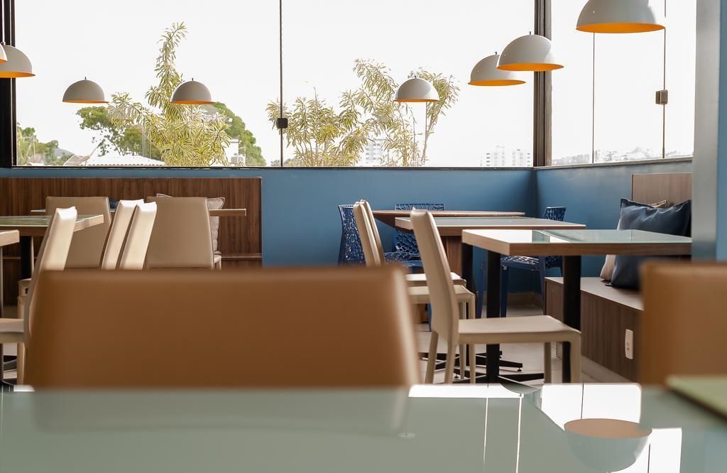 Um restaurante moderno com mesas e cadeiras de vidro, iluminação pendente no teto e vista para árvores e edifícios através de grandes janelas.