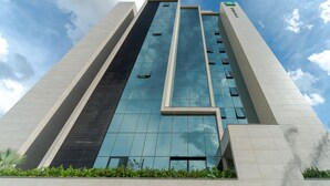 Un moderno edificio de gran altura con fachada de vidrio reflectante, un diseño vertical llamativo y un letrero verde en la parte superior.