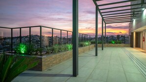 Terraza en la azotea con vista a la ciudad al atardecer, que cuenta con barandilla de cristal, plantas verdes y una estructura de pérgola.