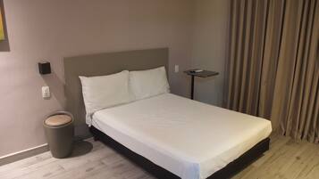 A neatly made bed in a hotel room with a wooden floor, a small bedside table, and a trash can.