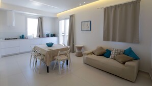 A modern living room with a beige sofa, a dining table with chairs, and a kitchen area with white cabinets and a window. - Appartamento Agamennone (Ragusa)