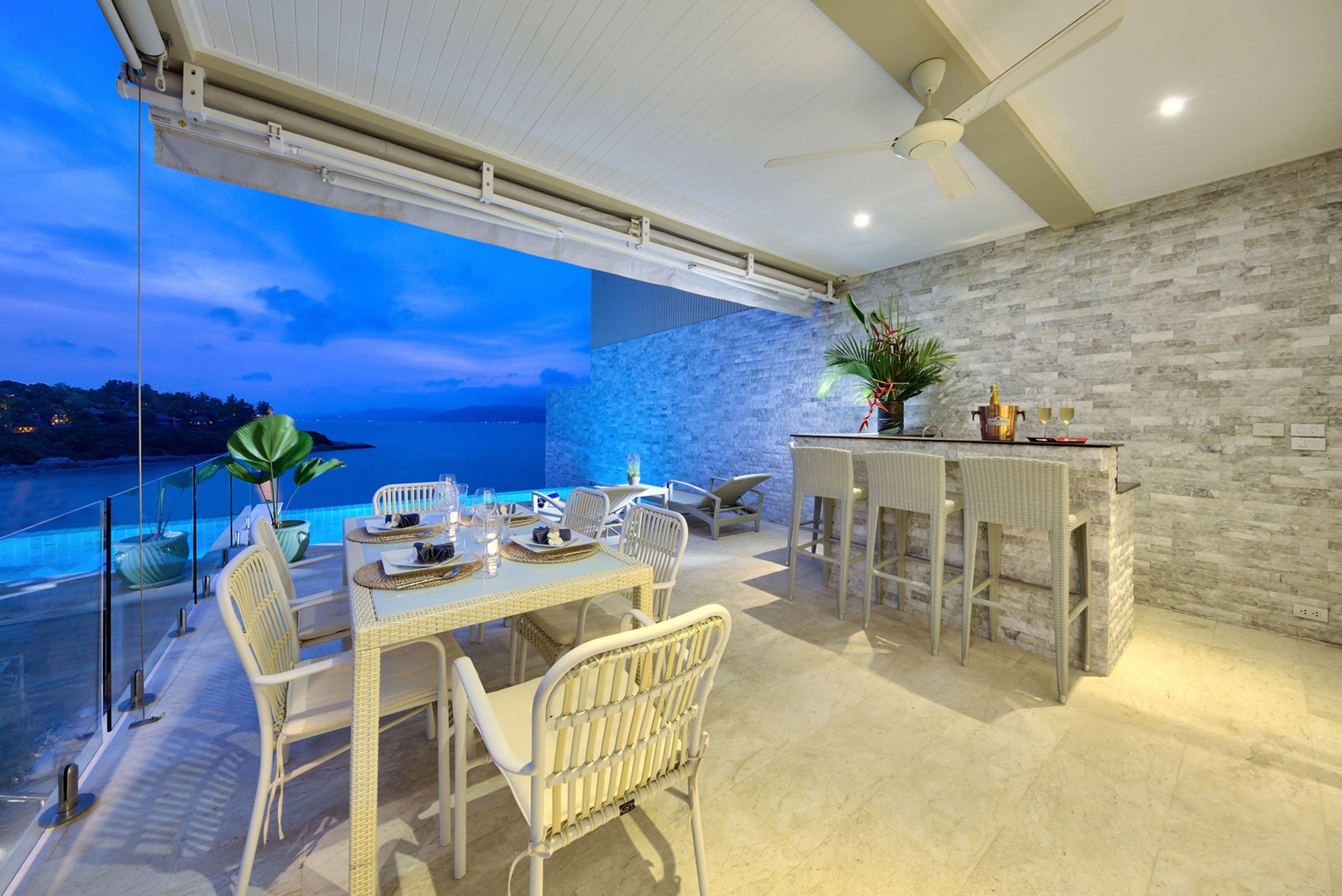A dining area with a view of the sea, a stone wall, and a bar with a plant.