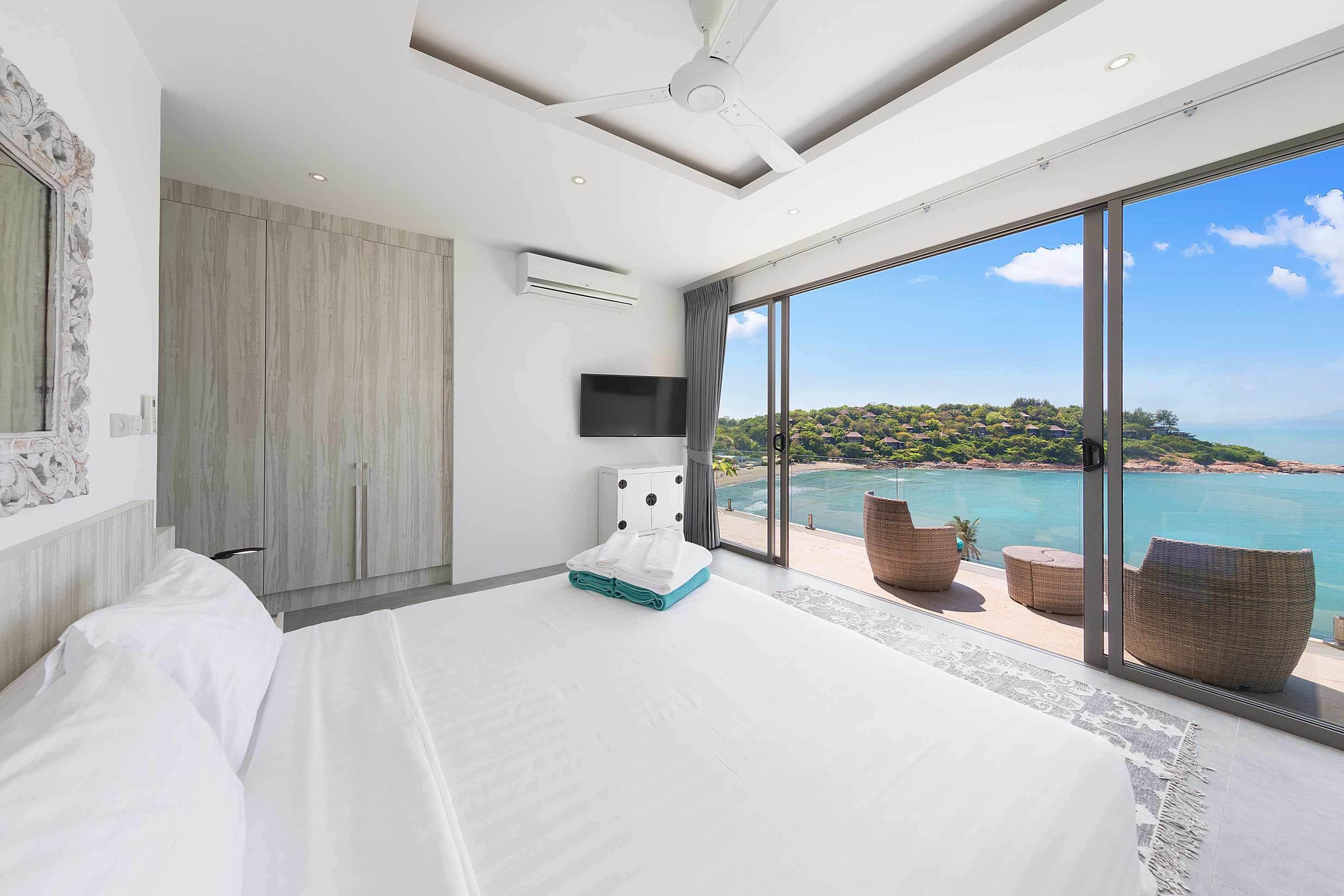 A modern hotel room with a large bed, a flat-screen TV, and a balcony with wicker furniture overlooking a coastal view.