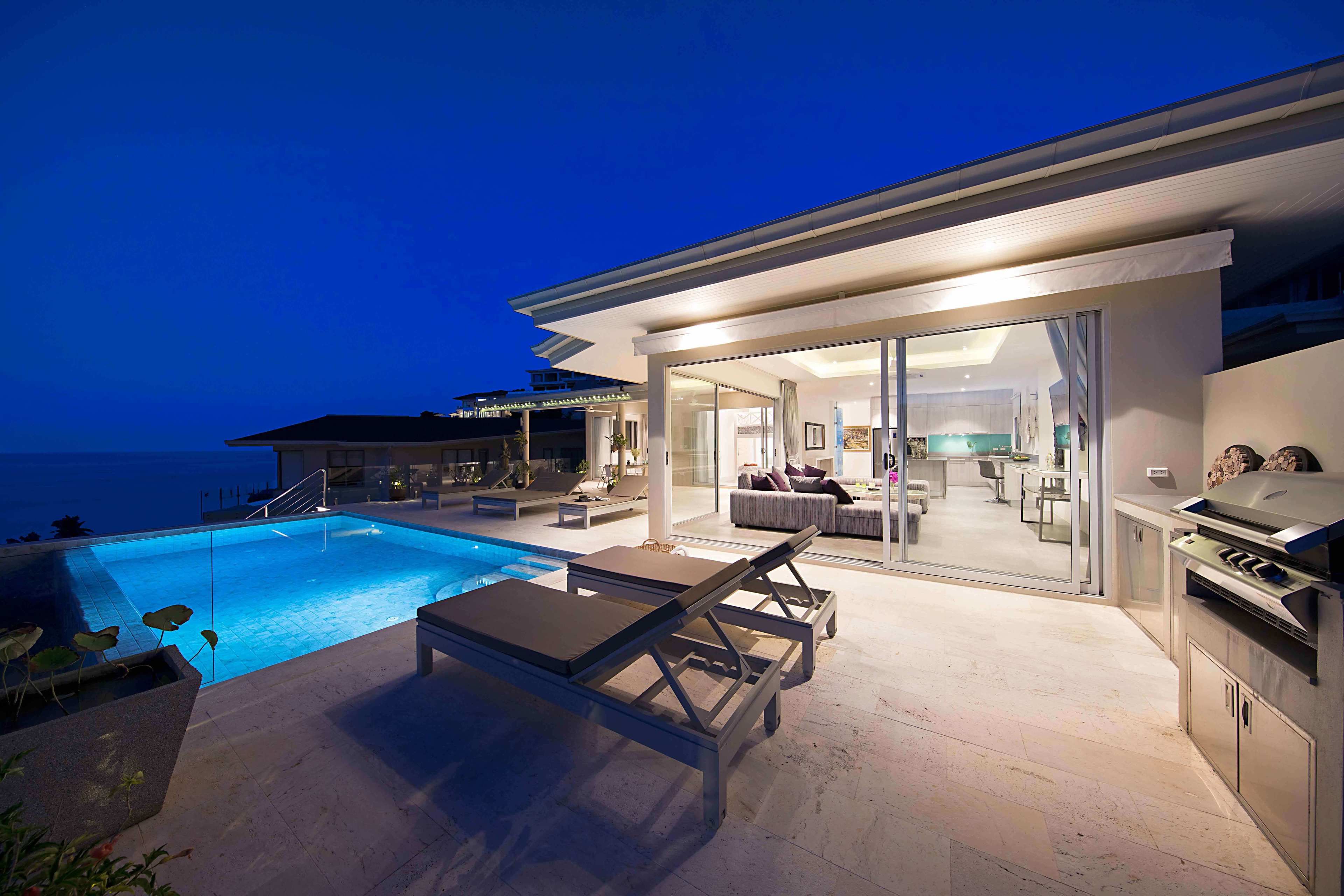 A modern outdoor pool area with lounge chairs, a barbecue grill, and a glass-enclosed living space with a view of the ocean.
